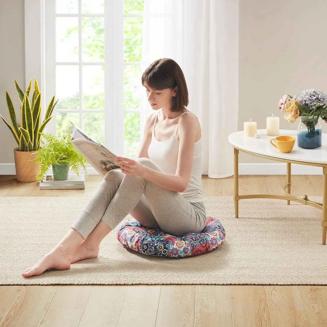 Woman comfortably reading in a bright room while seated on a round boho floor cushion, illustrating the pillow’s supportive design.
