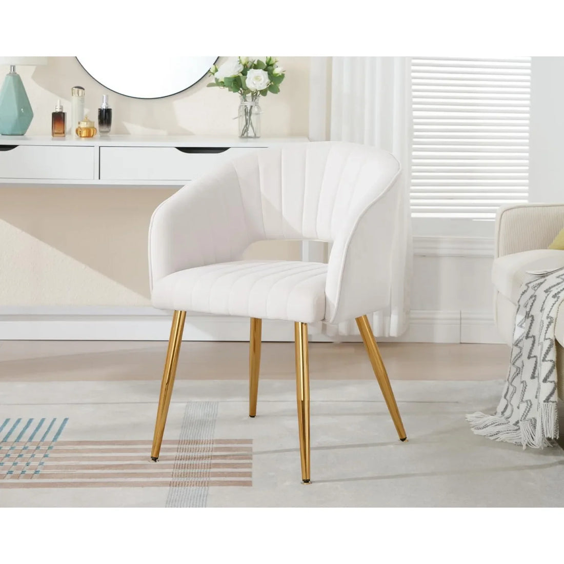 A cream velvet tufted vanity chair with a curved back and gold legs, placed next to a vanity table in a bright, modern bedroom setting.

